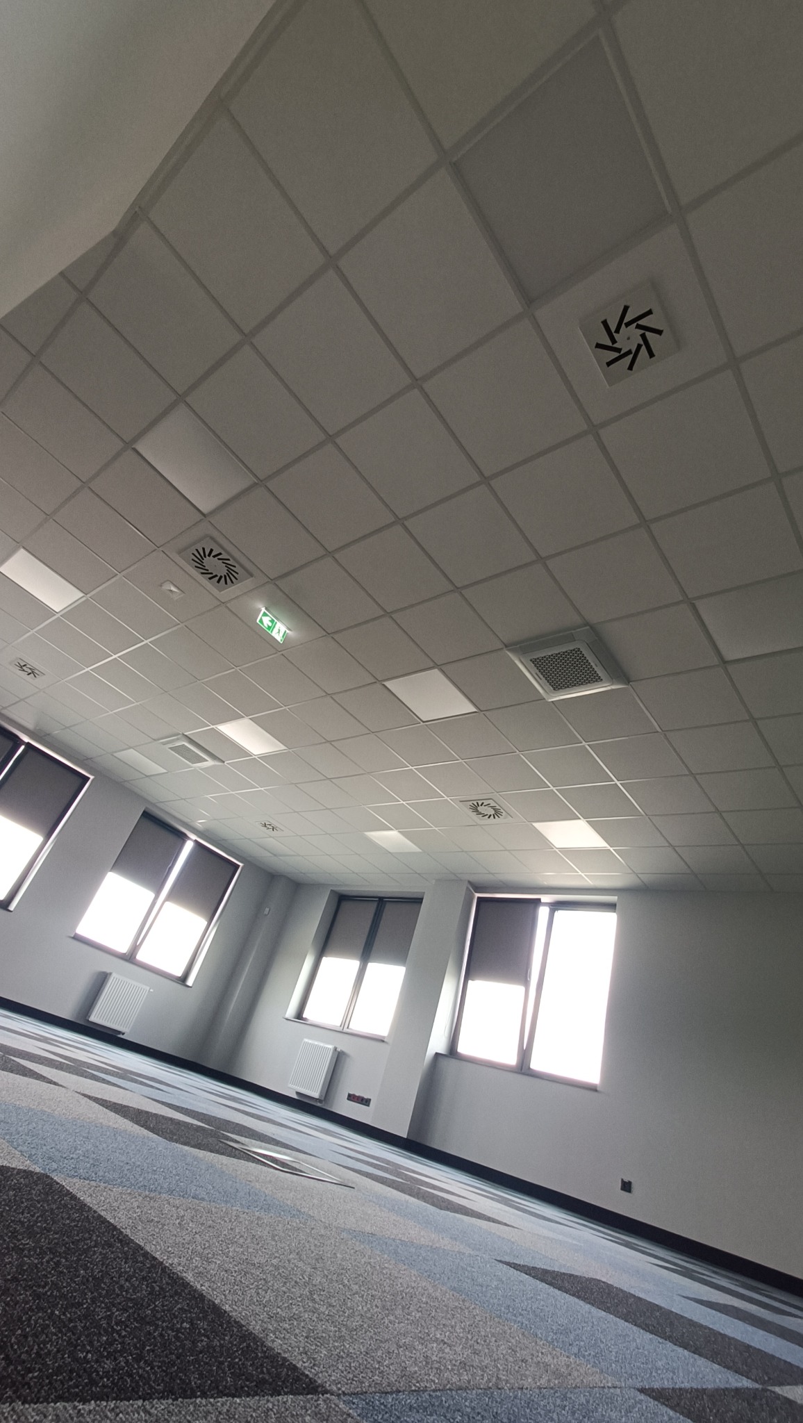 Wnętrze biurowe z kasetonowym sufitem podwieszanym, kratkami wentylacyjnymi, oknami z roletami i wzorzystą wykładziną podłogową.
