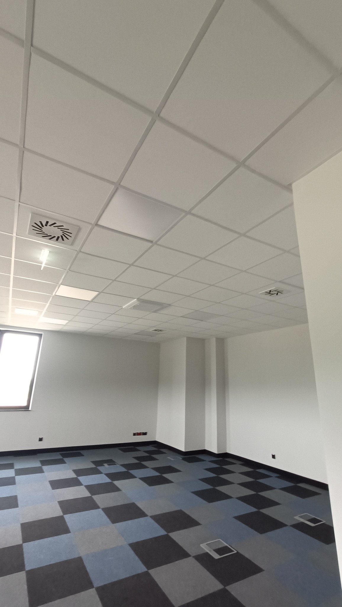 Białe panele sufitowe w biurze z kratką wentylacyjną, wzorzysta podłoga w szaro-niebieską szachownicę, widok na jasne okno.