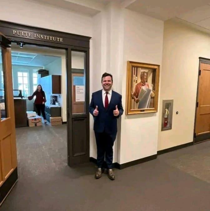 Uśmiechnięty mężczyzna w garniturze z czerwonym krawatem stoi w korytarzu biurowym przed obrazem i wejściem do Pulte Institute, pokazując dwa kciuki w górę.