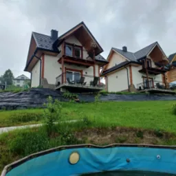 Dwa identyczne domy szkieletowe z drewnianymi balkonami i czarnymi dachami, widoczne z perspektywy basenu, na zboczu wzgórza.