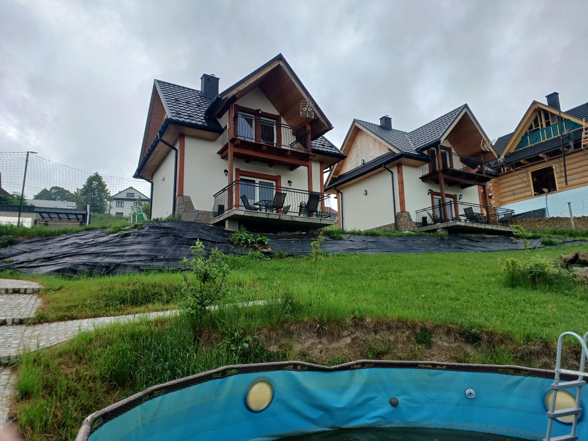 Dwa identyczne domy szkieletowe z drewnianymi balkonami i czarnymi dachami, widoczne z perspektywy basenu, na zboczu wzgórza.