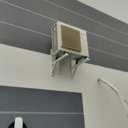 Zewnętrzna jednostka klimatyzacji Panasonic zamontowana na białej ścianie budynku z szarymi pasami, widoczne przewody i uchwyty montażowe.