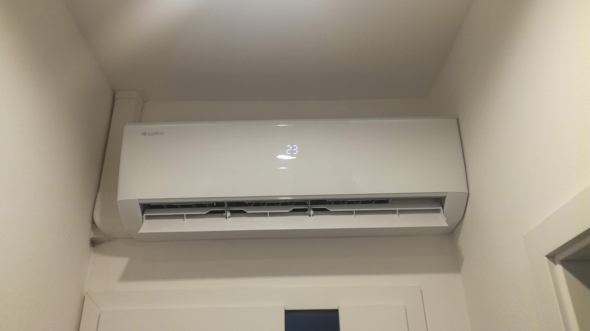 Biały klimatyzator marki Gree zamontowany na ścianie nad drzwiami, wyświetlający temperaturę 23 stopnie Celsjusza.