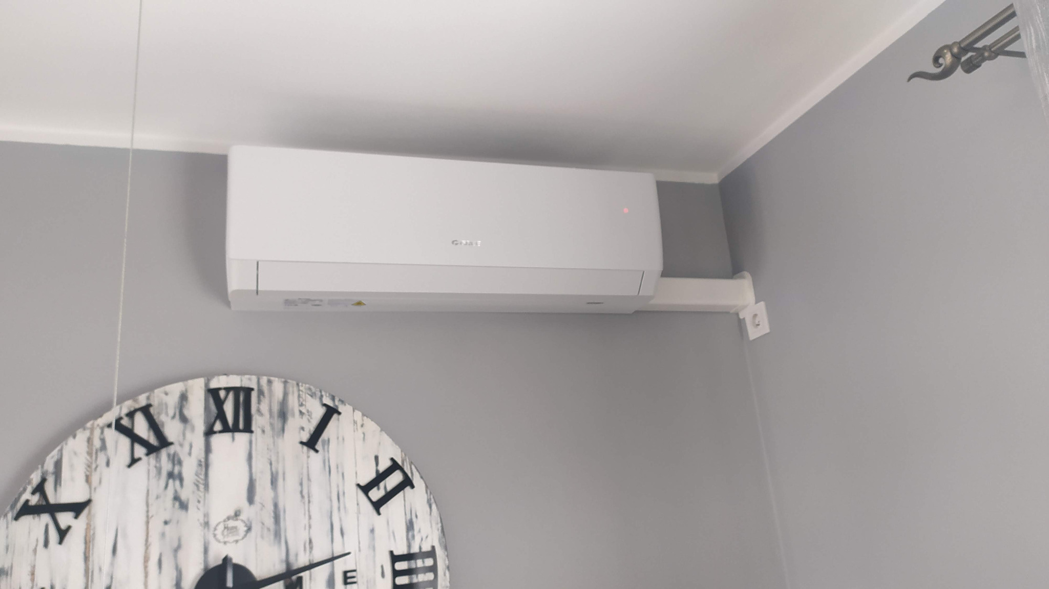 Biała klimatyzacja marki Gree zamontowana na szarej ścianie, poniżej widoczny ozdobny zegar w stylu vintage.