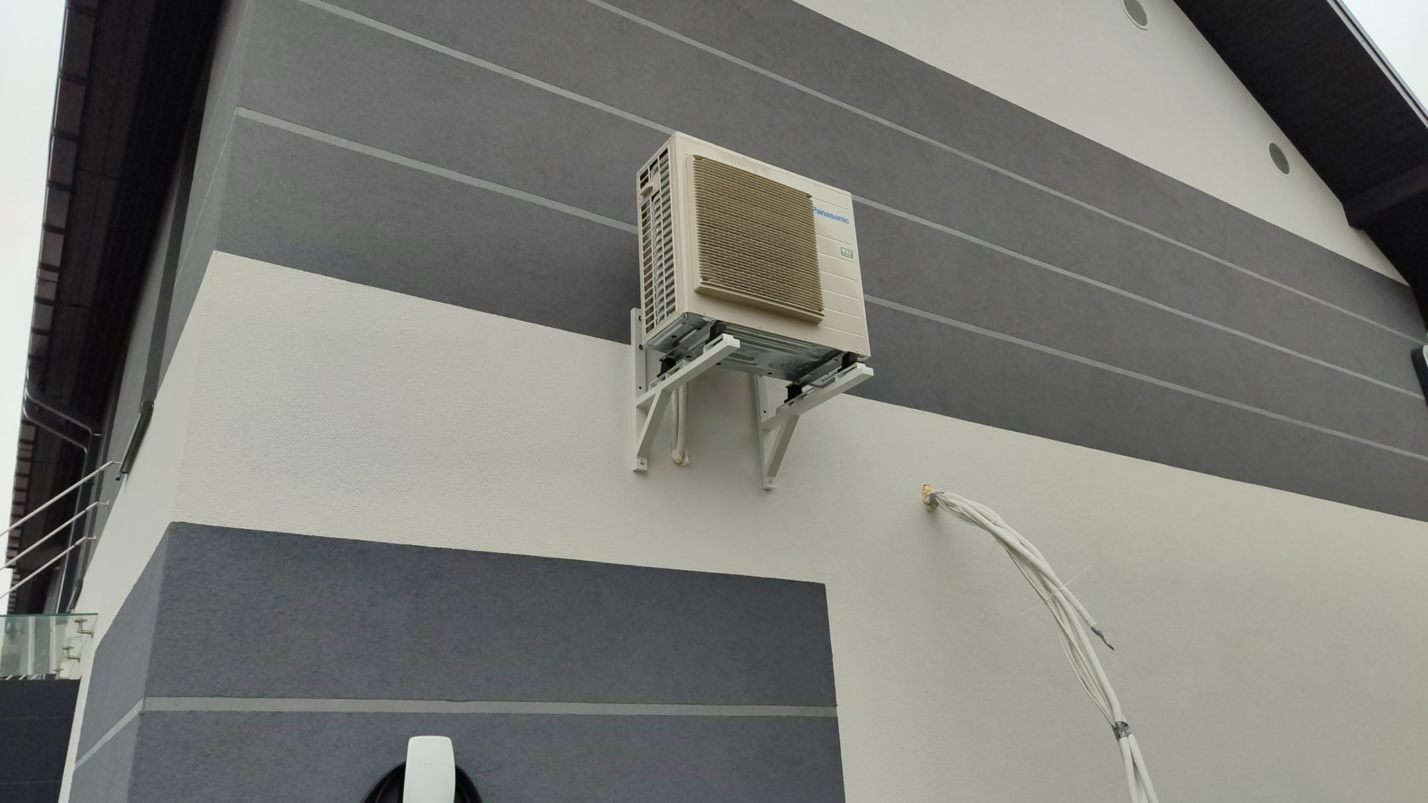 Zewnętrzna jednostka klimatyzacji Panasonic zamontowana na białej ścianie budynku z szarymi pasami, widoczne przewody i uchwyty montażowe.
