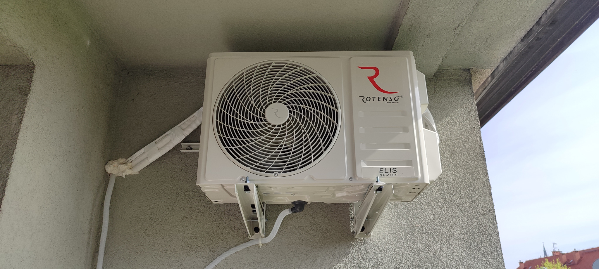 Zewnętrzna jednostka klimatyzacji Rotenso Elis Series zamontowana na szarej elewacji budynku, widoczne orurowanie z izolacją termiczną.