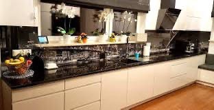 Biała, nowoczesna kuchnia z czarnym blatem i panelem szklanym z motywem miejskim nad blatem, widoczne kwiaty w doniczkach, owoce w koszyku i czarny okap.