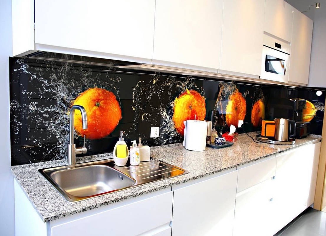 Szklany panel kuchenny z motywem pomarańczy i efektami rozpryskującej się wody, zamontowany nad blatem z szarego granitu i białymi szafkami kuchennymi.