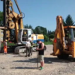 Praca geodezyjna na placu budowy: operator niwelatora z tyczką, w tle wiertnica palowa na gąsienicach i koparko-ładowarka JCB, pracownicy w kamizelkach odblaskowych.