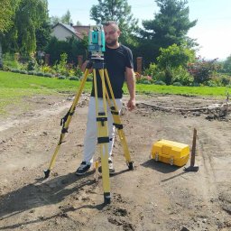 SYGNATA GEODEZJA - Mężczyzna w czarnej koszulce obsługuje niwelator na trójnogu w terenie przygotowanym pod budowę, obok żółta skrzynka narzędziowa i wbity kołek.