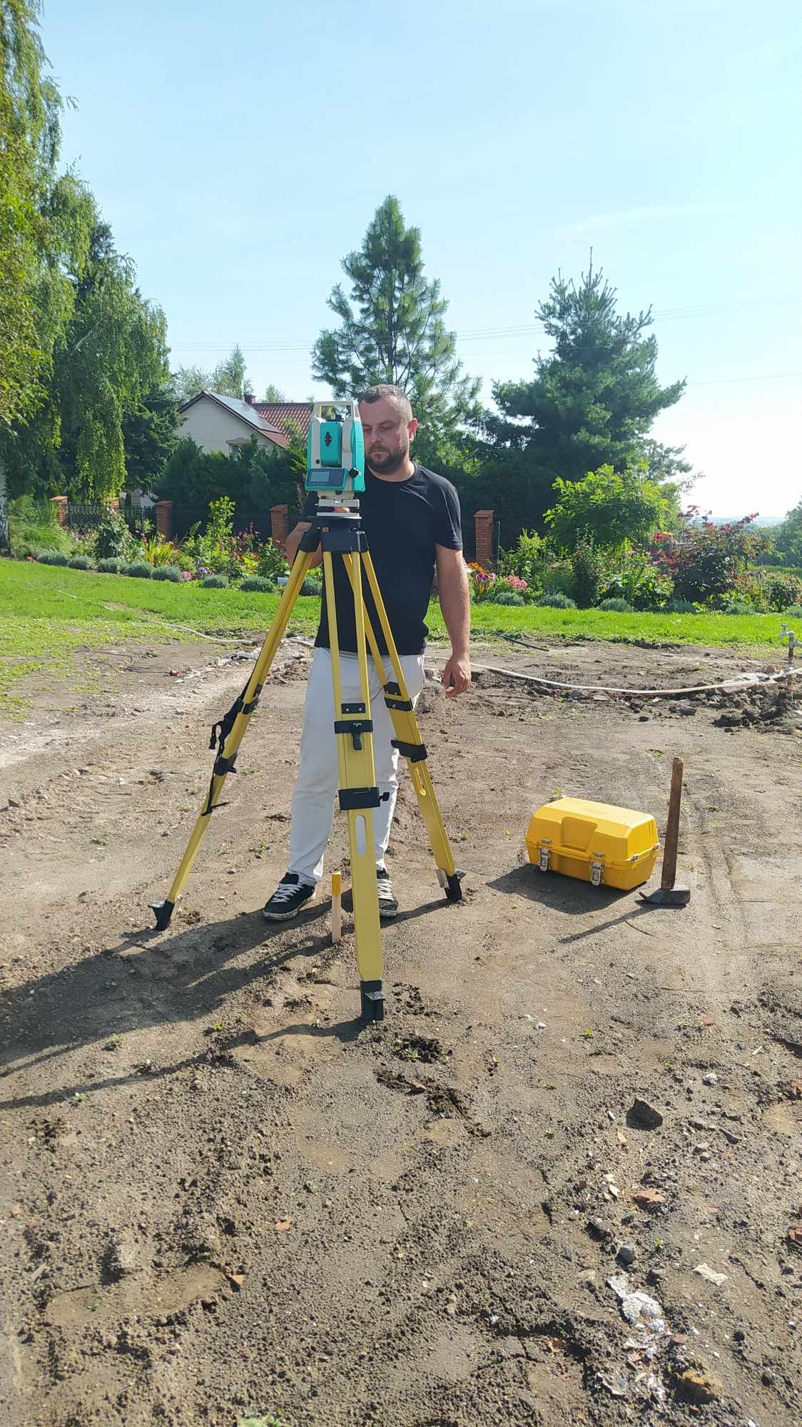 Mężczyzna w czarnej koszulce obsługuje niwelator na trójnogu w terenie przygotowanym pod budowę, obok żółta skrzynka narzędziowa i wbity kołek.