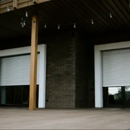 Dwa okna z częściowo opuszczonymi, szarymi roletami zewnętrznymi, umieszczone w ciemnej, ceglanej elewacji budynku z drewnianym tarasem i dekoracyjnymi żarówkami.