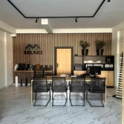 Wnętrze biura z drewnianą ścianą z listew, jasnym biurkiem, czterema czarnymi krzesłami i marmurową podłogą. Widoczny fragment logo firmy SULARZ na ścianie.