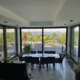 Nowoczesne wnętrze jadalni z dużymi oknami przesłoniętymi poziomymi żaluzjami fasadowymi, widok na zieleń za oknem, marmurowa podłoga, czarne krzesła i stół.