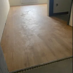 Ułożona, jasnobrązowa wykładzina imitująca drewno w pustym pokoju z grzejnikiem pod oknem, widoczna listwa przypodłogowa i próg drzwiowy.