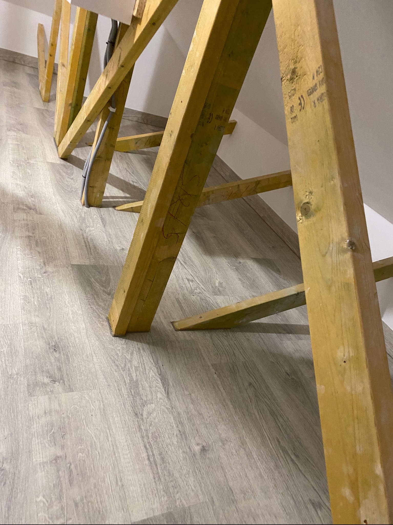 Drewniane rusztowanie pod sufitem na tle jasnej, panelowej podłogi, widoczne przewody elektryczne.