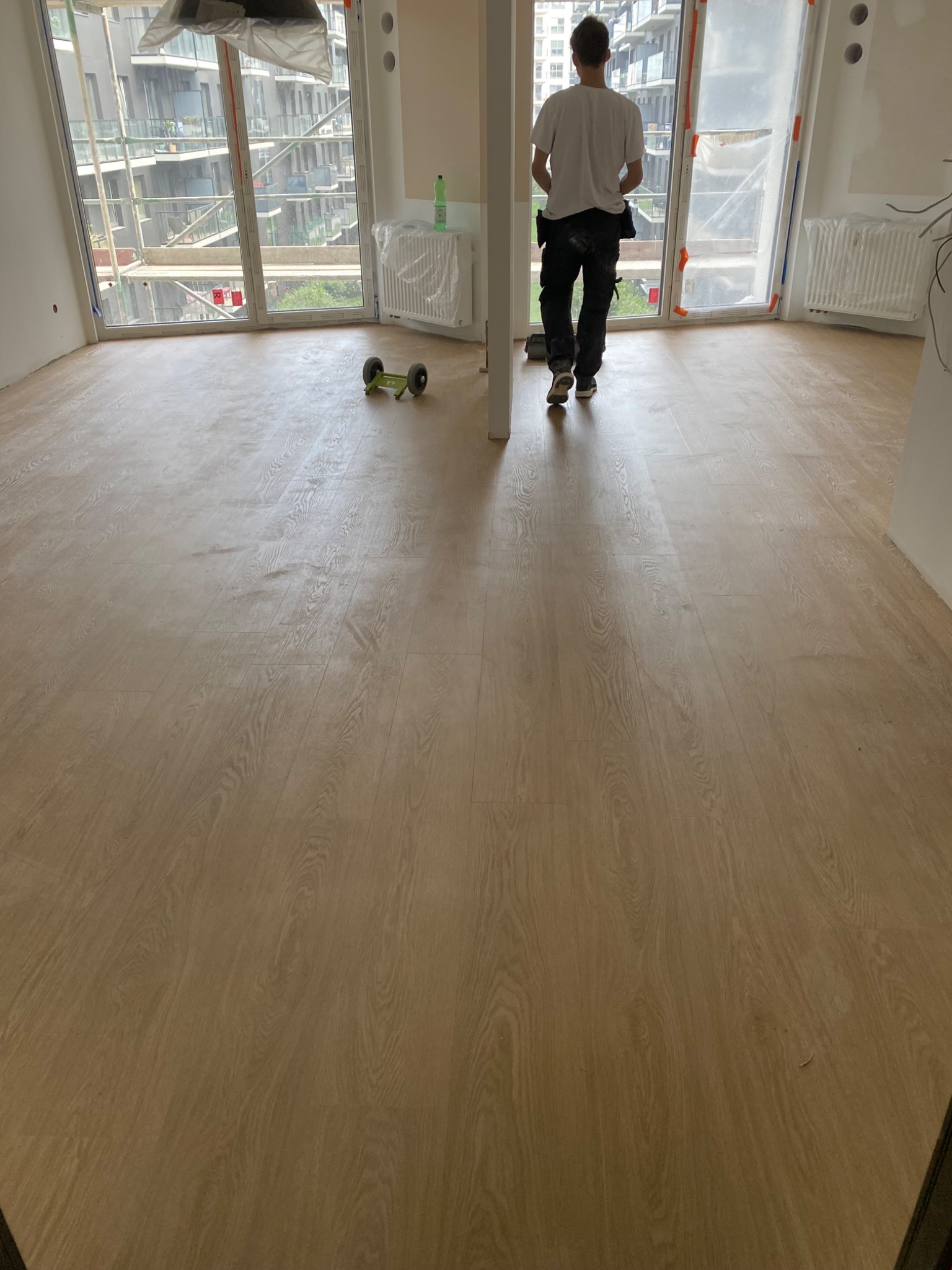 Jasna, drewnopodobna podłoga w nowym mieszkaniu, widok na okna zabezpieczone folią, pracownik w oddali z narzędziami.