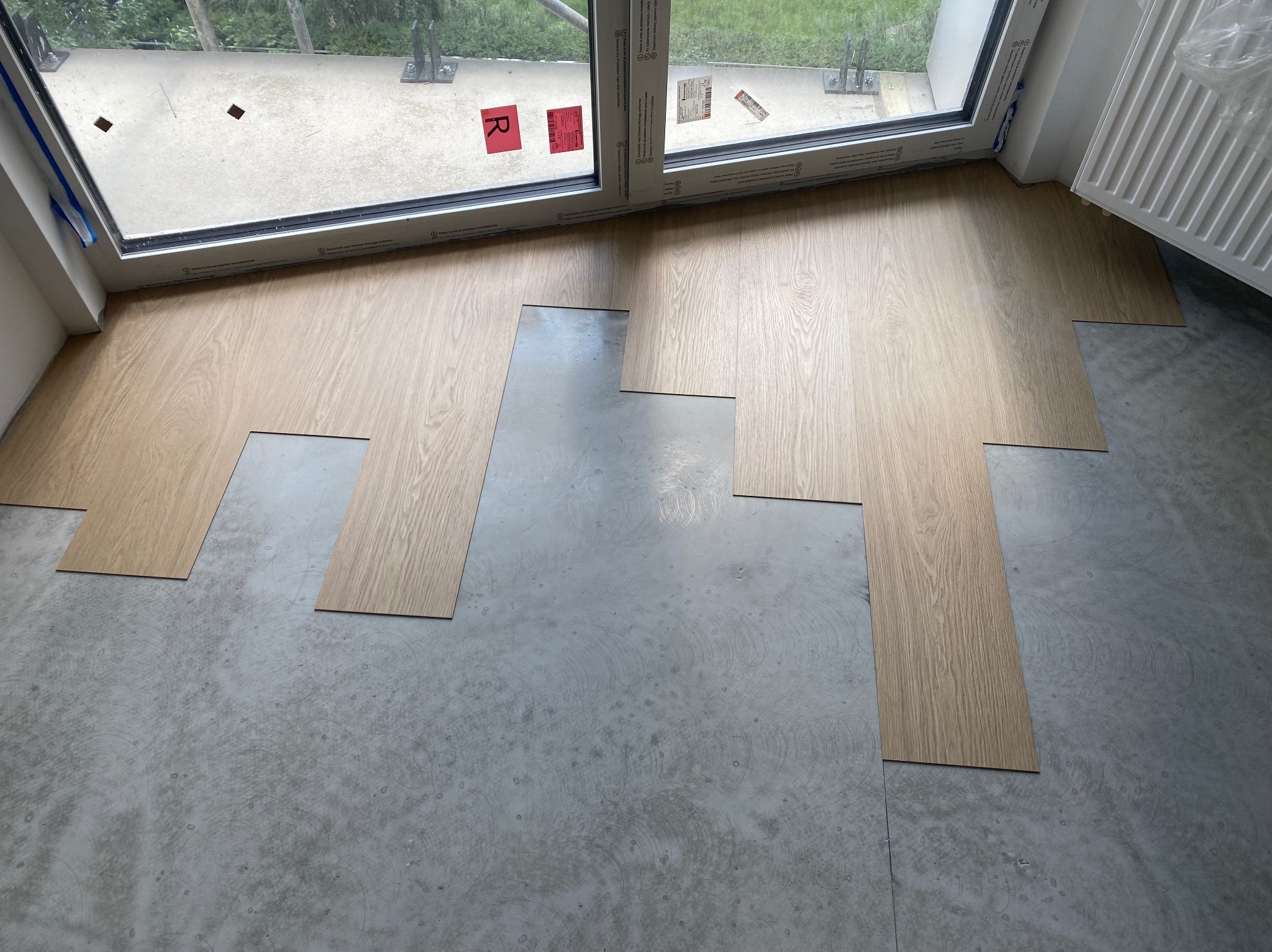 Układanie paneli podłogowych w surowym wnętrzu, widoczne fragmenty podłogi bez wykończenia, okno z naklejkami i grzejnik.