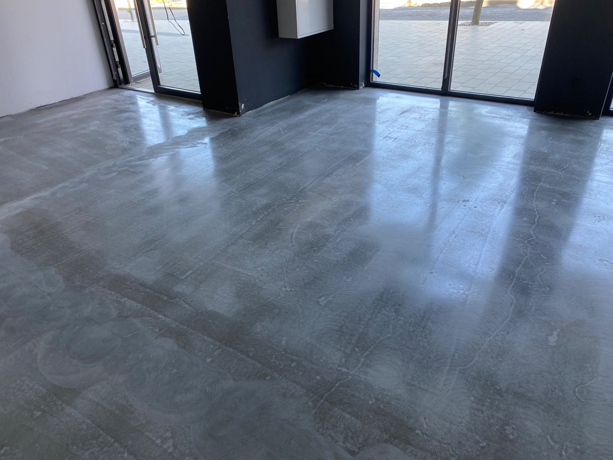 Świeżo wylana, mokra posadzka betonowa w jasnym pomieszczeniu z dużymi oknami, widoczne ślady po rozprowadzaniu masy.