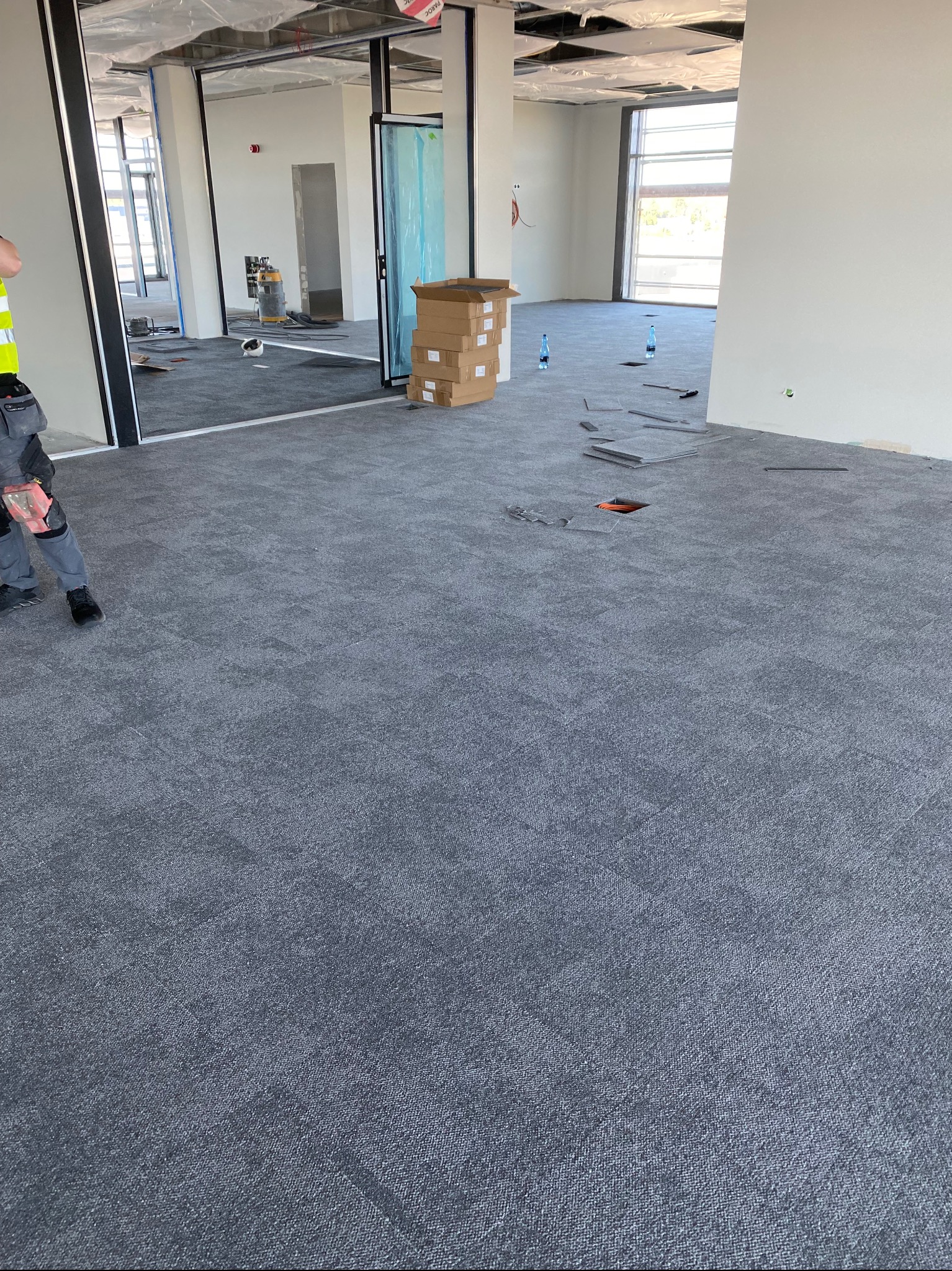 Montaż szarej wykładziny dywanowej w płytkach w biurze, widoczne kartony z płytkami, rozłożone narzędzia i fragment pracownika.