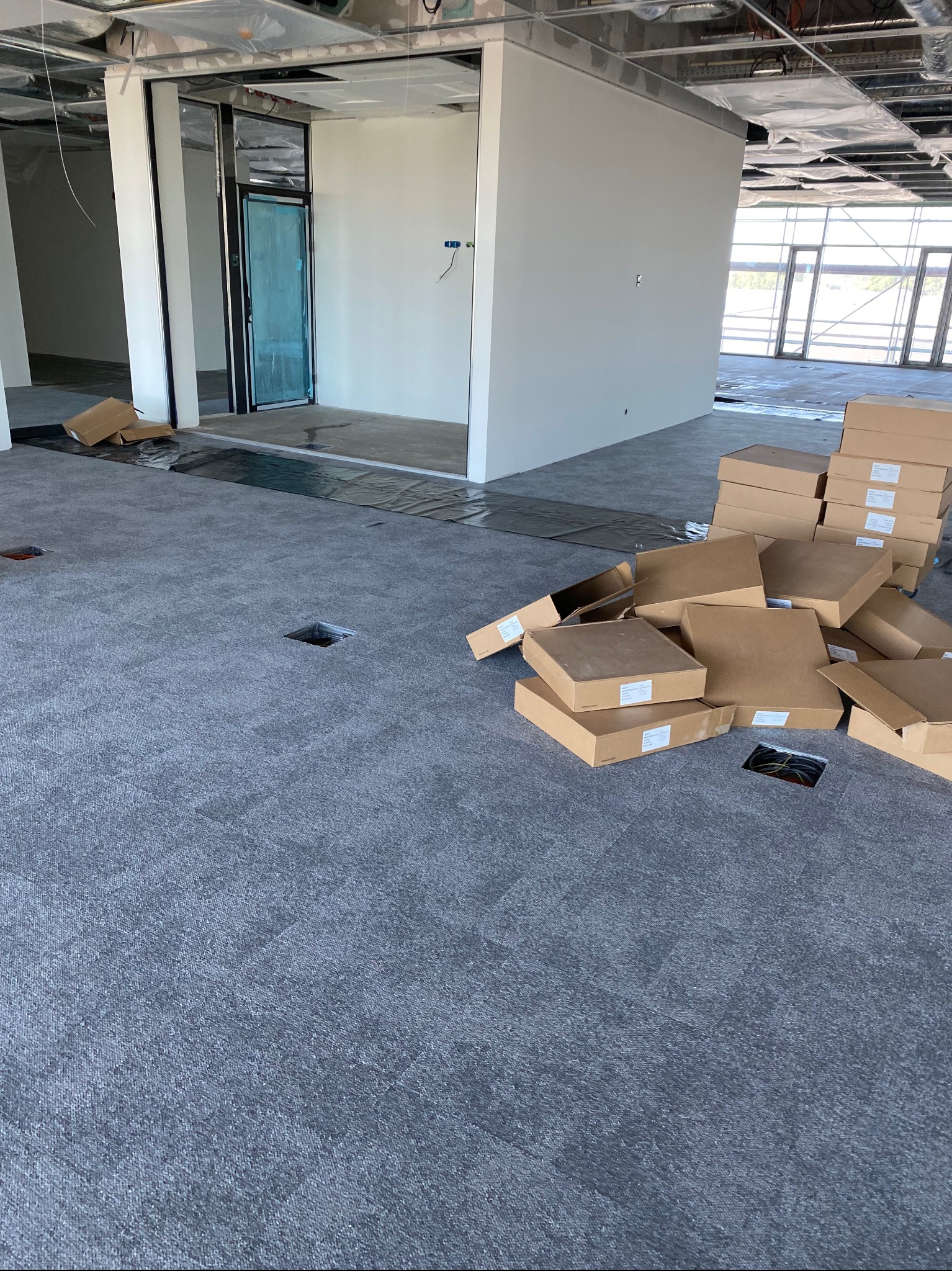 Szara wykładzina dywanowa w nowym biurze z otworami na gniazdka i stertą kartonów z nową wykładziną w tle.