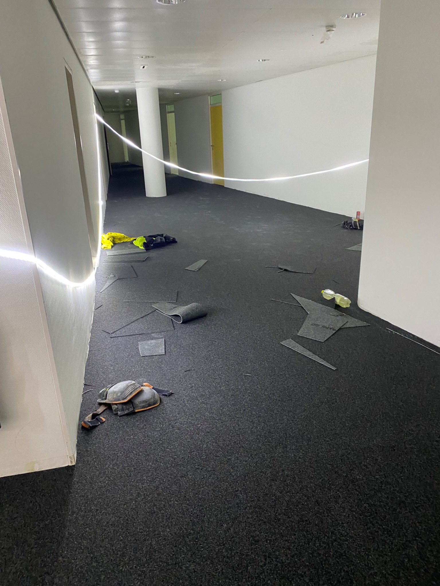 Korytarz w trakcie układania ciemnej wykładziny dywanowej w płytkach, widoczne narzędzia, fragmenty dociętych płytek oraz ochraniacze na kolana.