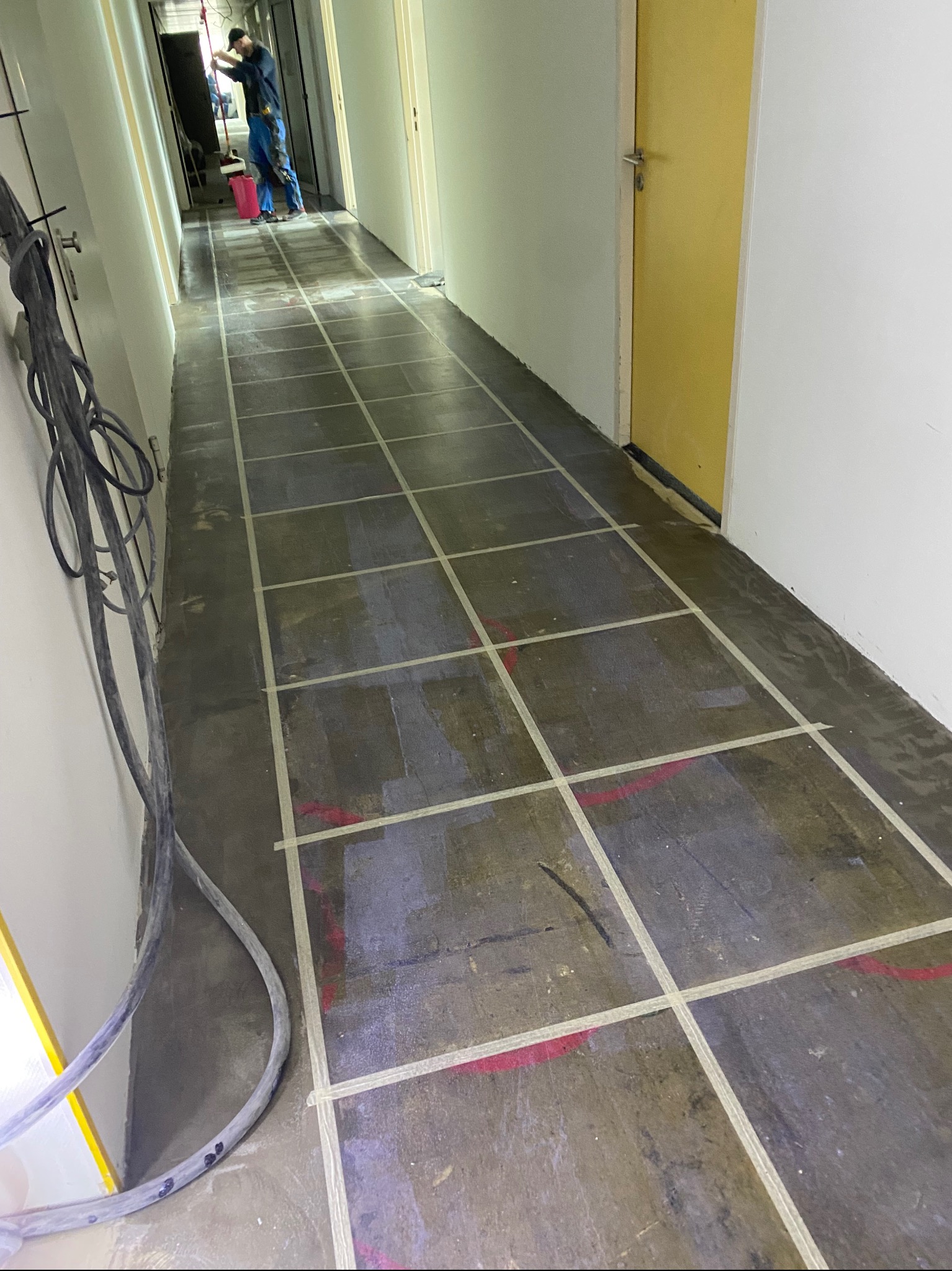 Korytarz w trakcie przygotowywania do położenia nowej podłogi, widoczne taśmy wyznaczające pola i pracownik w oddali z mopem i wiadrem.