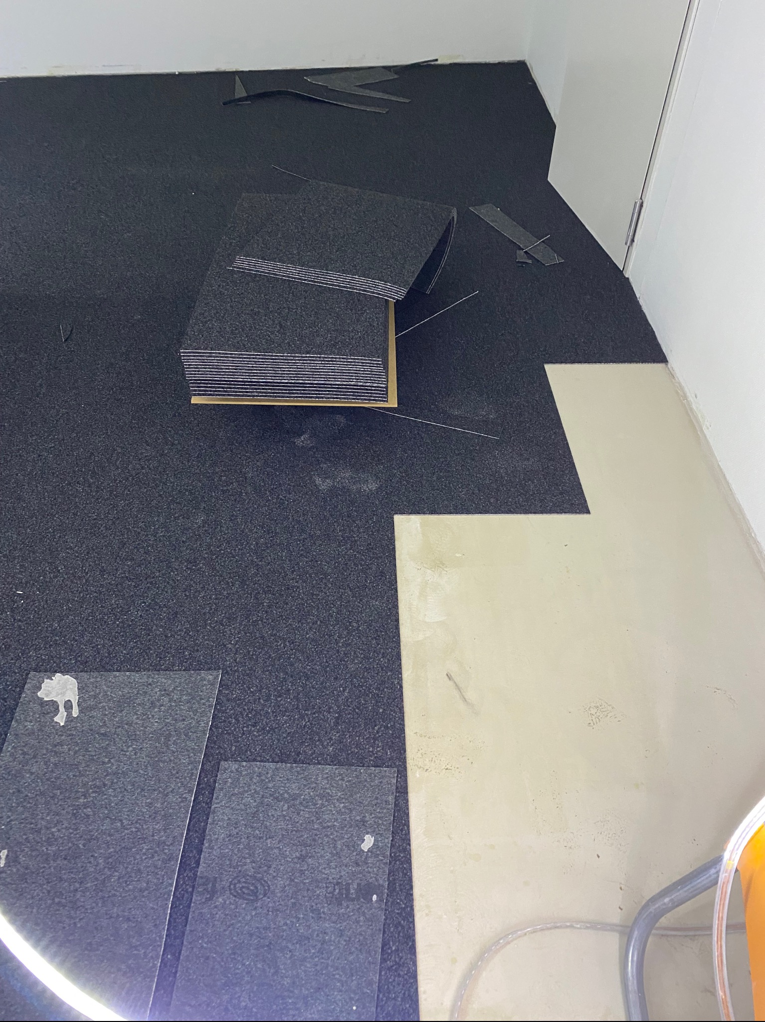 Układanie ciemnej wykładziny dywanowej w pomieszczeniu, widoczne docięte fragmenty i stos płytek, podłoże częściowo przygotowane.