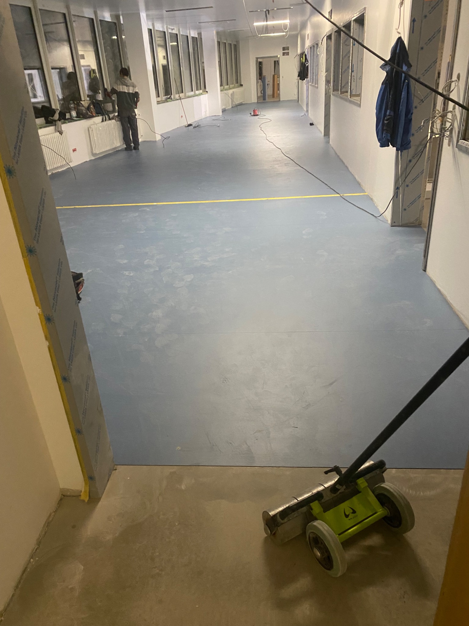 Długi korytarz w trakcie wykańczania, z położoną niebieską podłogą i żółtą linią, widoczny pracownik przy oknie oraz maszyna do dociskania wykładzin na pierwszym planie, surowy stan wykończenia...