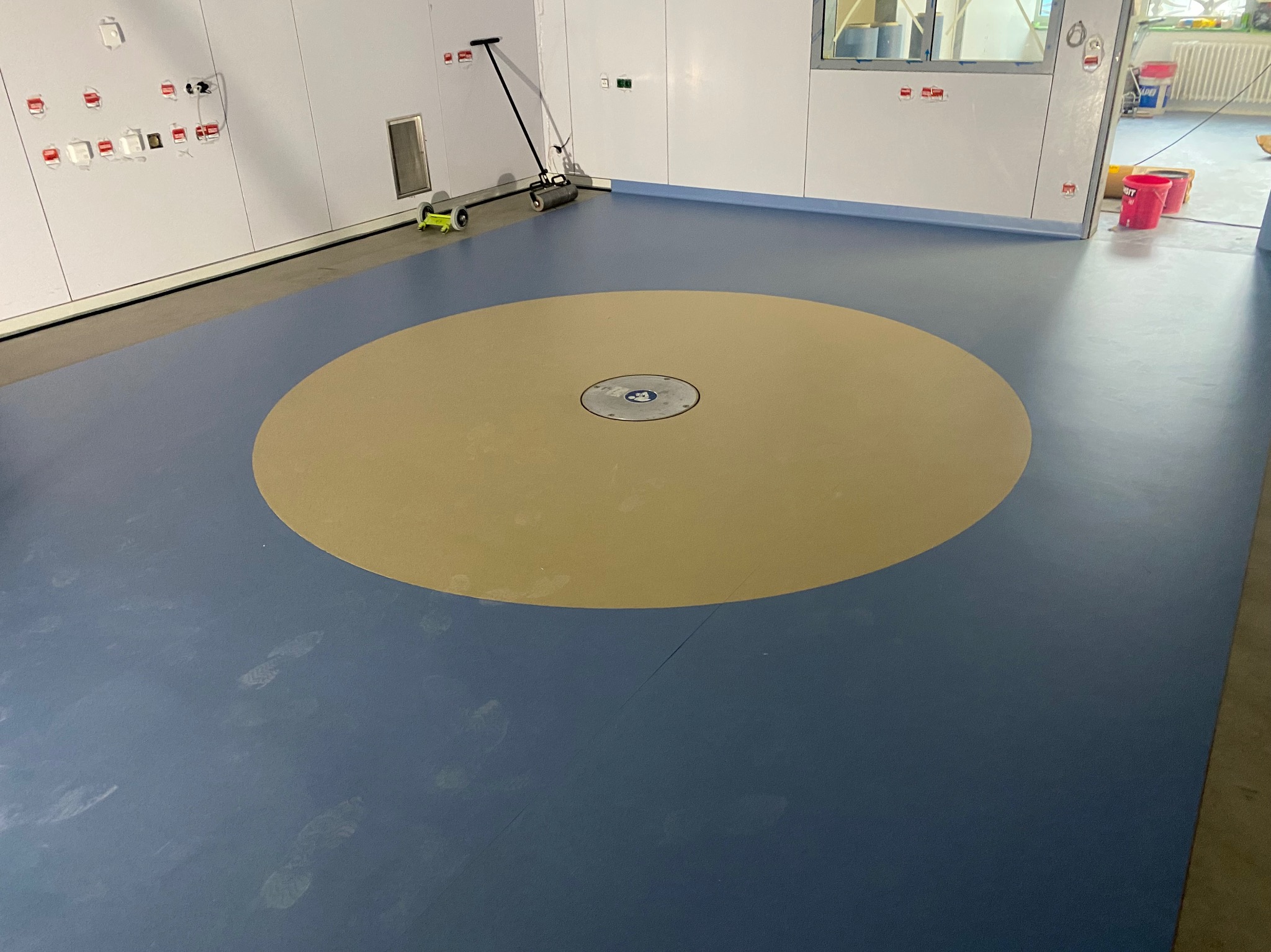 Niebiesko-beżowa wykładzina podłogowa z okrągłą strefą centralną i widocznymi śladami stóp, uzupełniona narzędziem do montażu wykładzin w tle, na tle białych ścian.