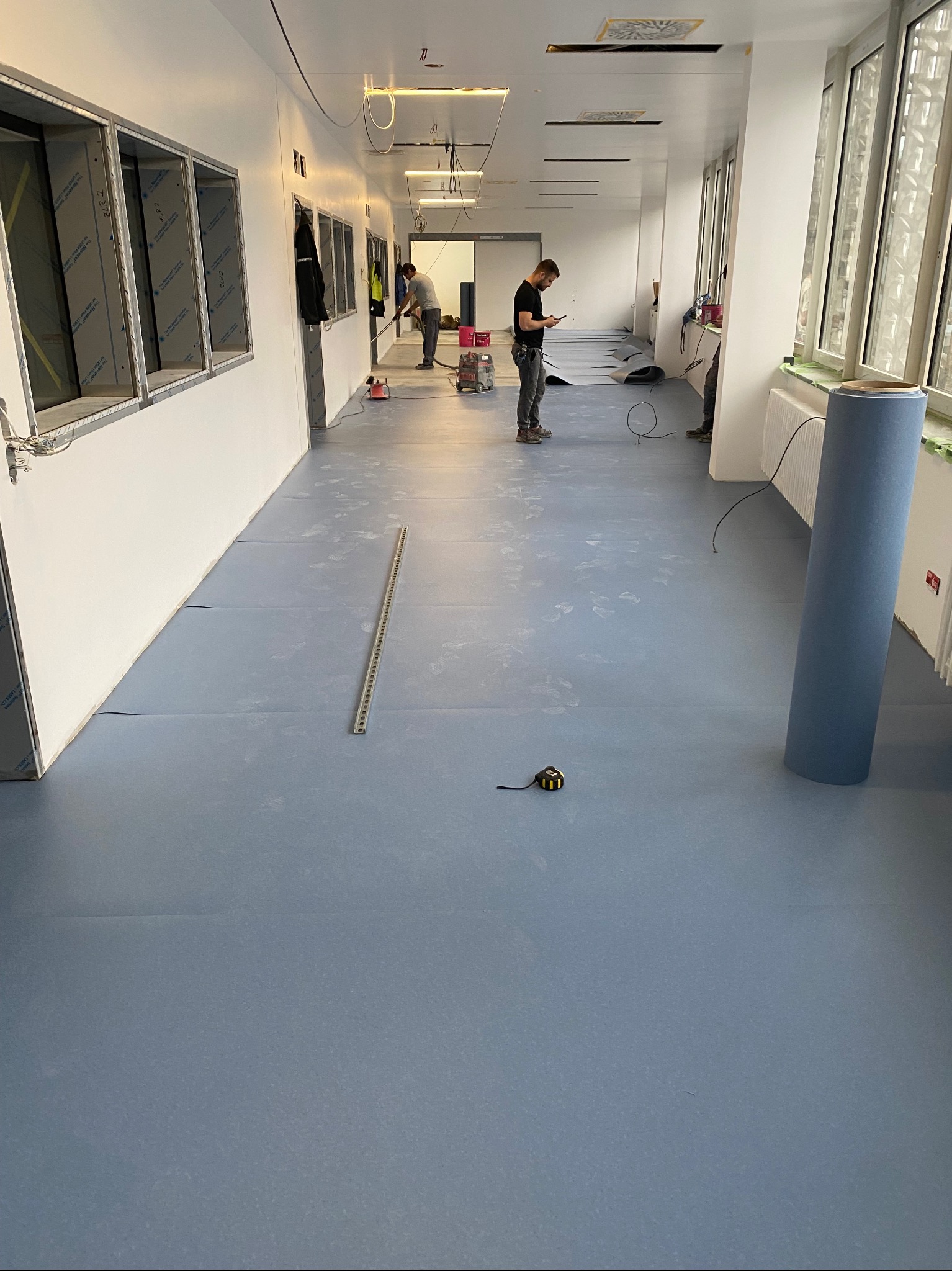 Montaż niebieskiej wykładziny w długim korytarzu z oknami, widoczny zwinięty rulon, miarka i pracownicy podczas instalacji.