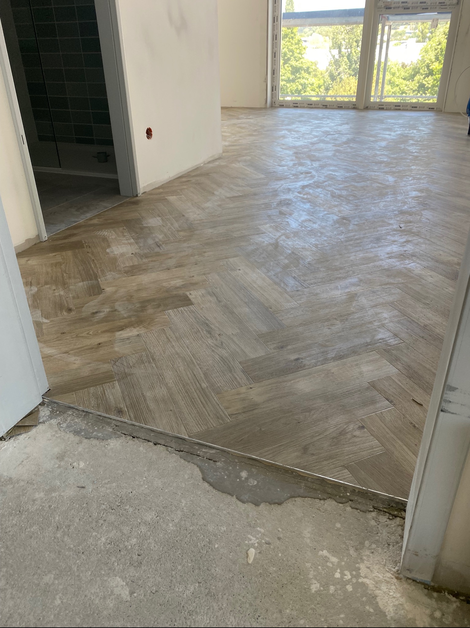 Ułożona w jodełkę jasna podłoga z paneli imitujących drewno, widok na fragment łazienki z ciemnymi płytkami i okno z widokiem na zieleń, surowy beton przy progu.