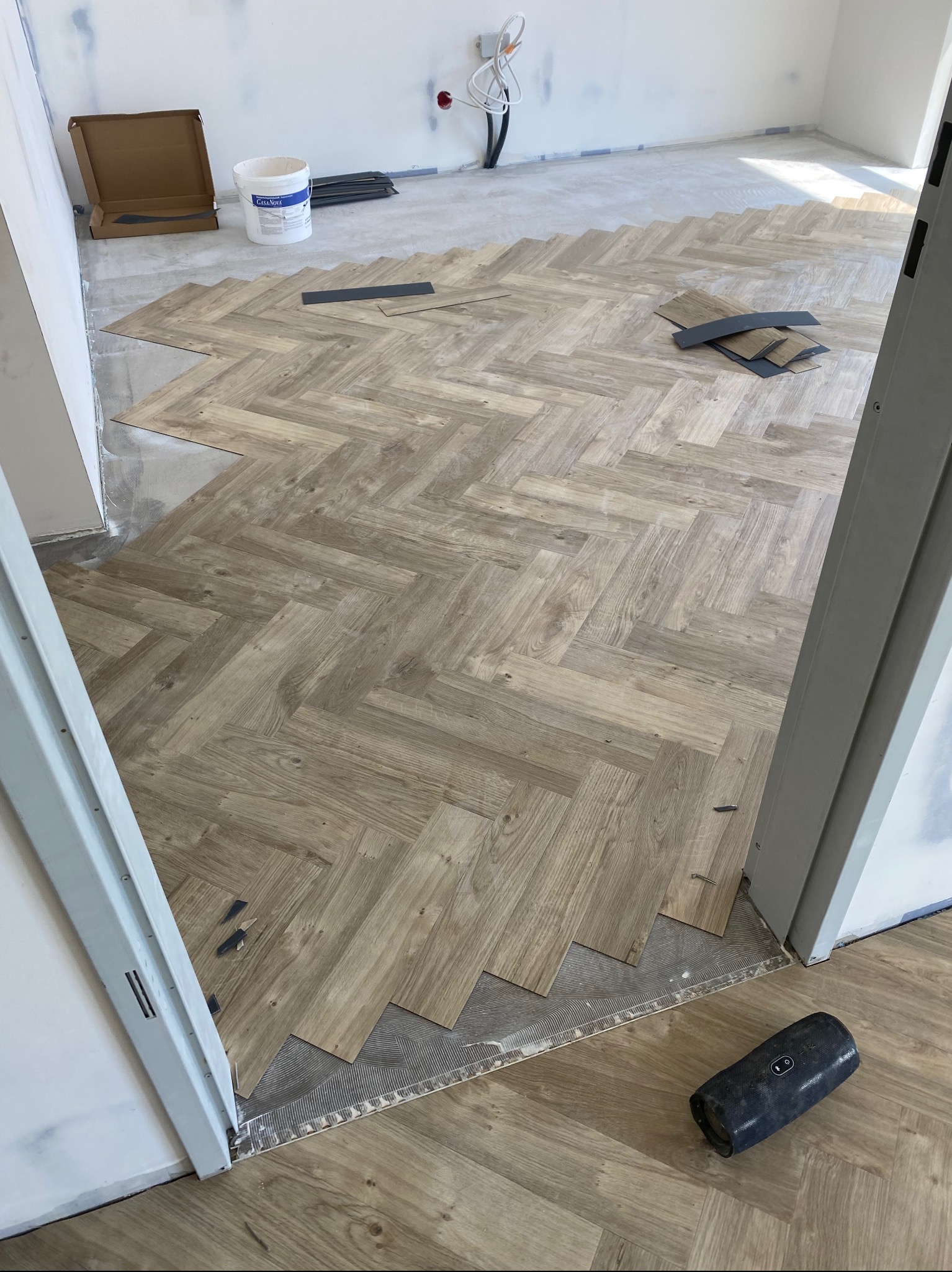 Układanie jasnej wykładziny w jodełkę, widoczny klej, docięte elementy i głośnik przenośny leżący obok framugi drzwi.