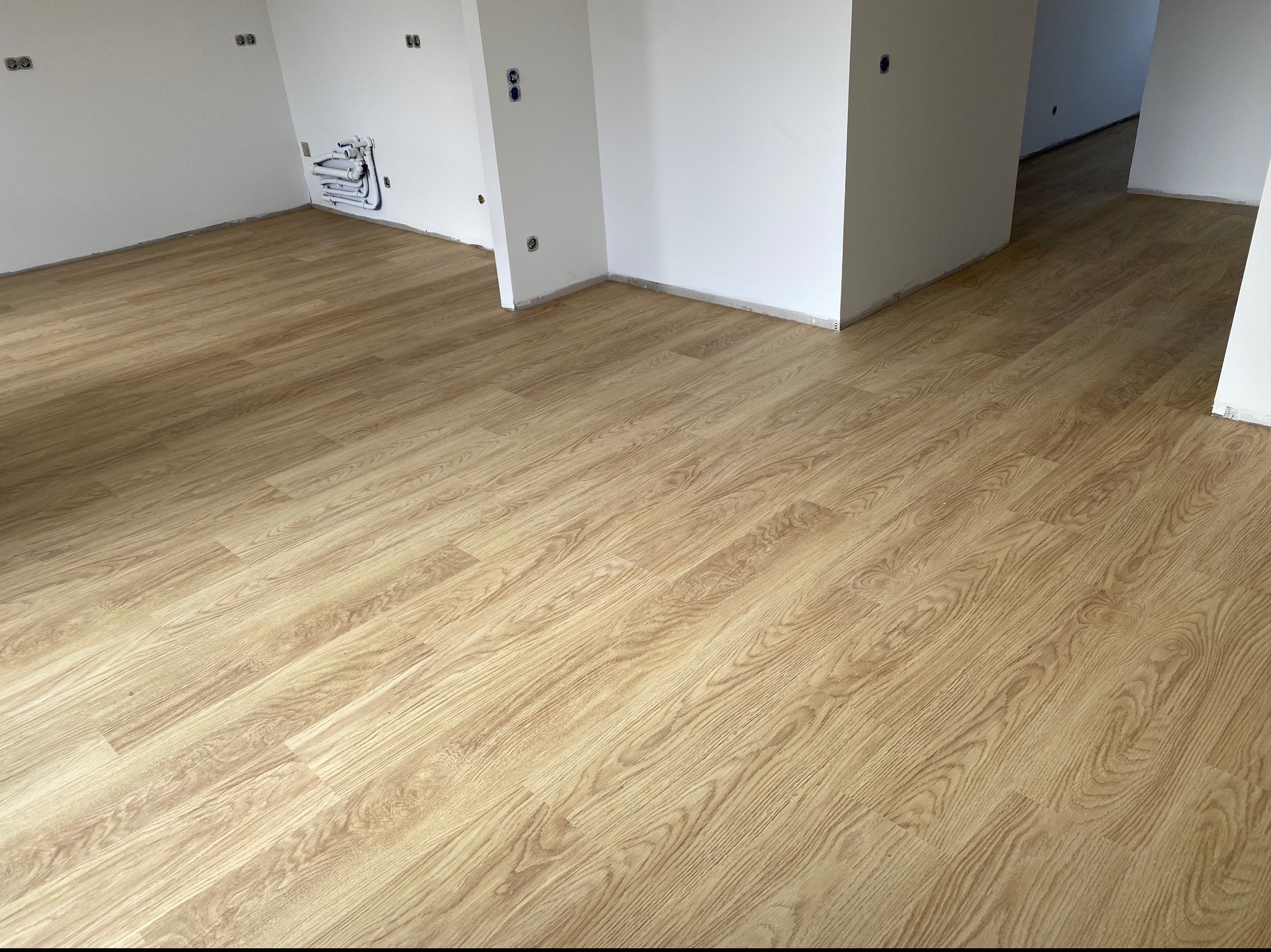 Jasna, drewnopodobna podłoga ułożona w nowym, pustym pomieszczeniu z białymi ścianami i widocznymi instalacjami elektrycznymi i hydraulicznymi.