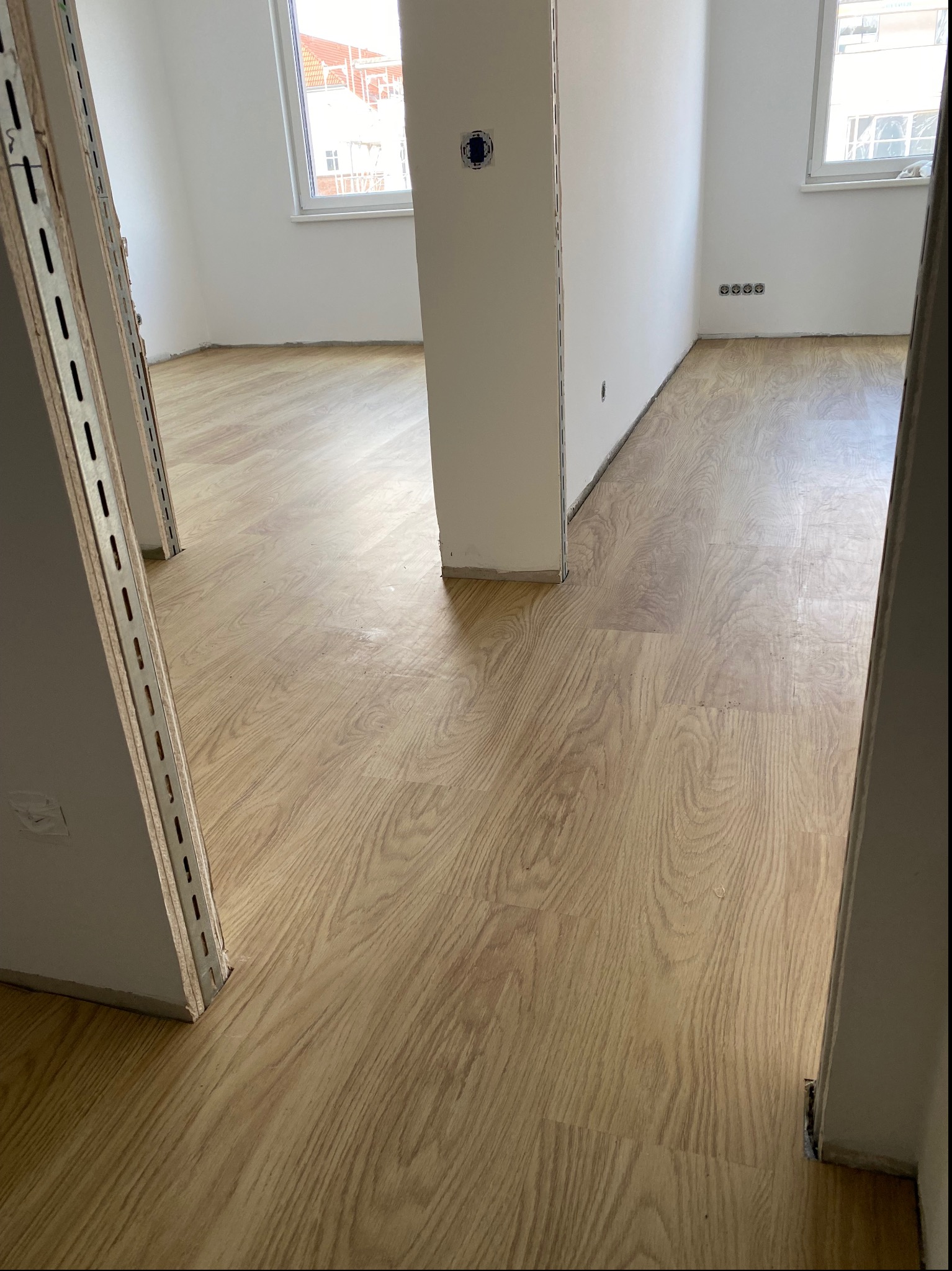 Jasna, drewnopodobna podłoga w trakcie montażu w nowym mieszkaniu, widoczne niedokończone ściany i metalowe profile przy futrynach.
