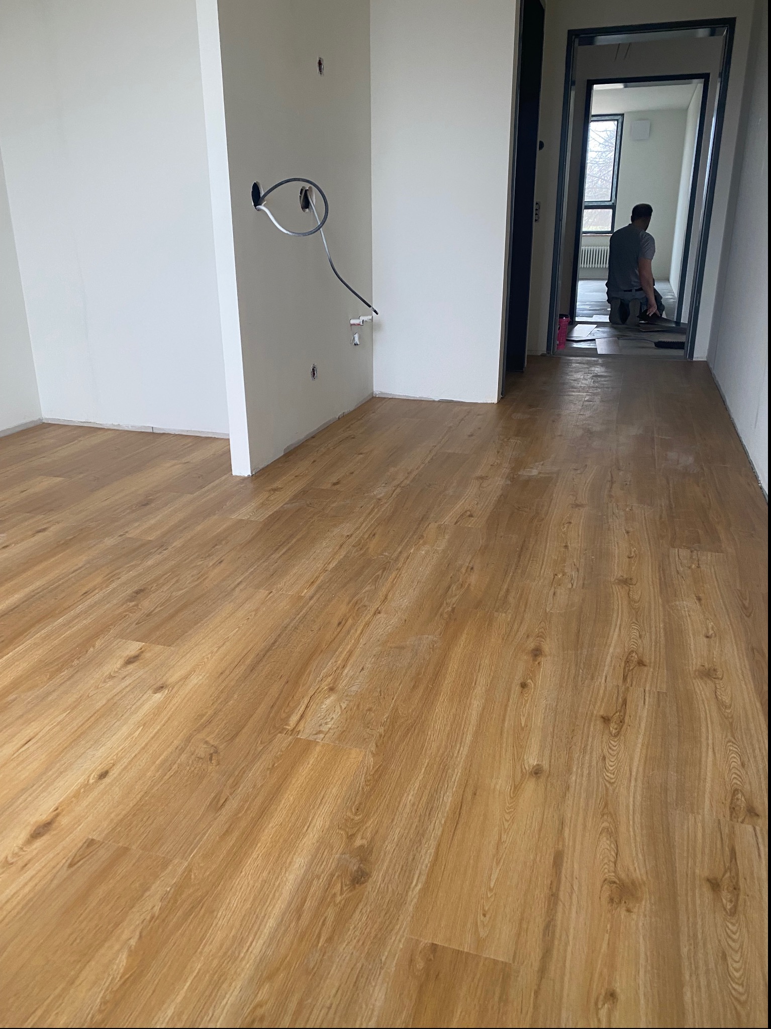 Układanie paneli podłogowych w jasnym pomieszczeniu, widoczne kable wystające ze ściany i pracownik w oddali.