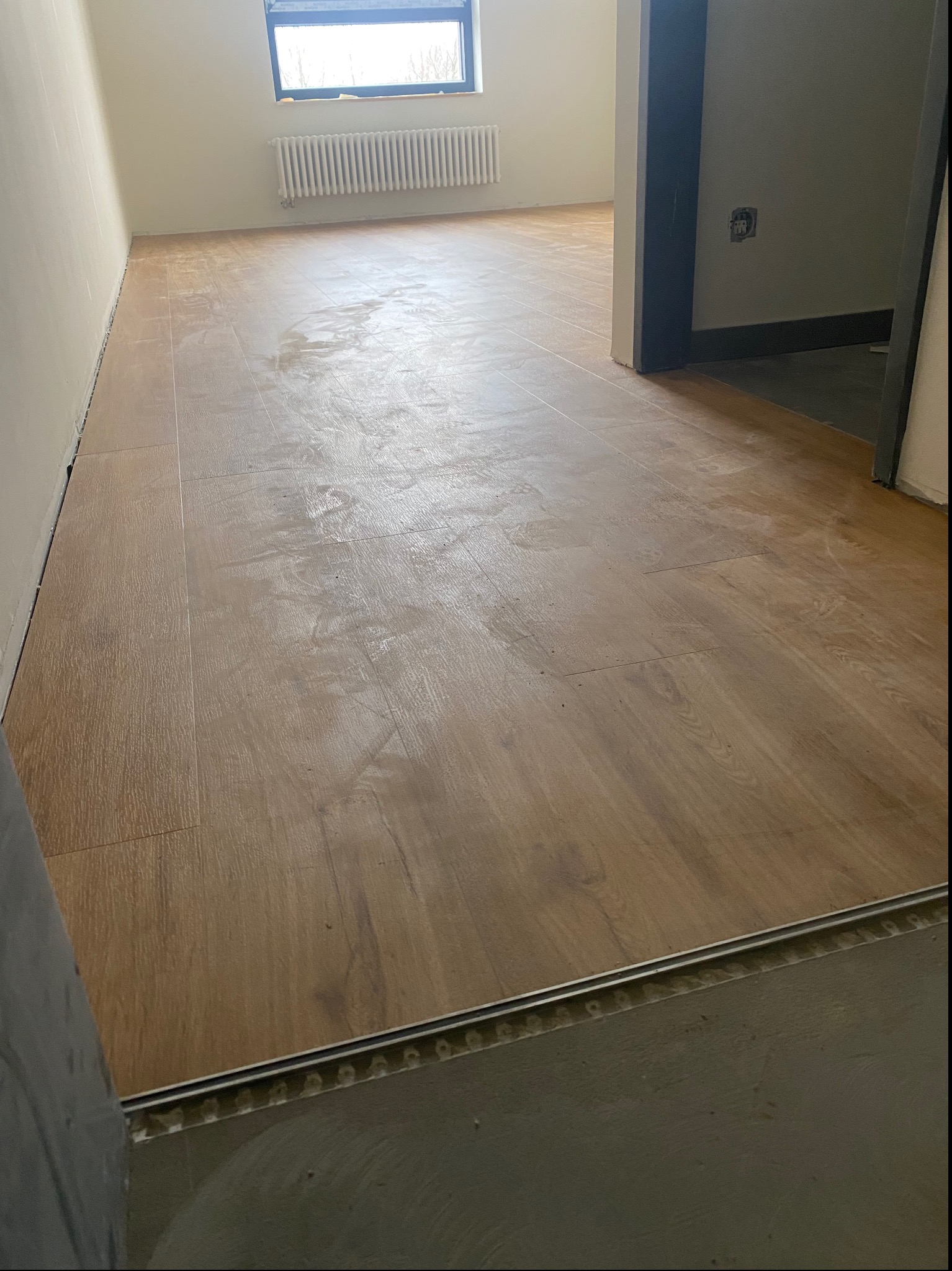 Ułożona, jasnobrązowa wykładzina imitująca drewno w pustym pokoju z grzejnikiem pod oknem, widoczna listwa przypodłogowa i próg drzwiowy.