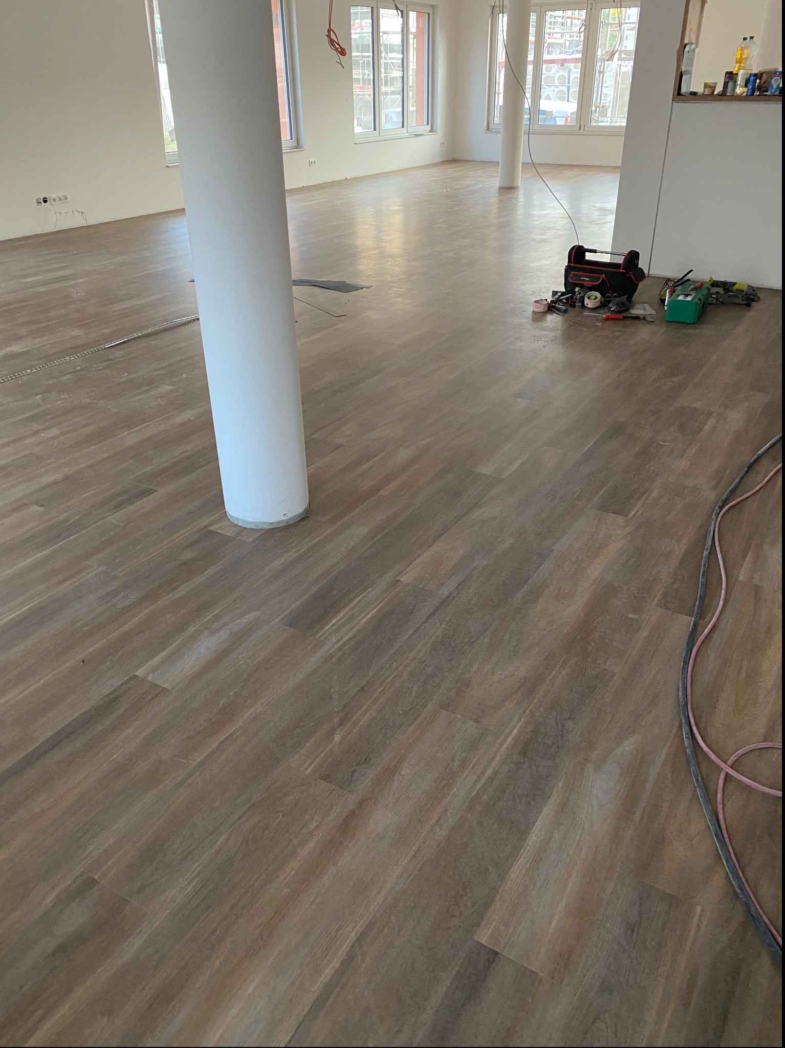 Układanie paneli podłogowych w przestronnym, jasnym pomieszczeniu z białymi kolumnami. Widoczne narzędzia i akcesoria do montażu podłogi. Częściowo wykończona podłoga z drewnopodobnych paneli.