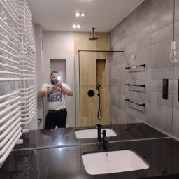 Budo-Wyko-Rem Bartłomiej Zmorzyński - Nowoczesna łazienka z podwójną umywalką na czarnym blacie, szare płytki na ścianach, drewniana kabina prysznicowa typu walk-in z czarną armaturą, grzejnik drabinkowy po lewej, odbicie osoby...