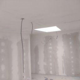 Budo-Wyko-Rem Bartłomiej Zmorzyński - Wnętrze w trakcie prac wykończeniowych: ściany pokryte gładzią, widoczne kable wystające z sufitu, przygotowane pod instalację oświetlenia.