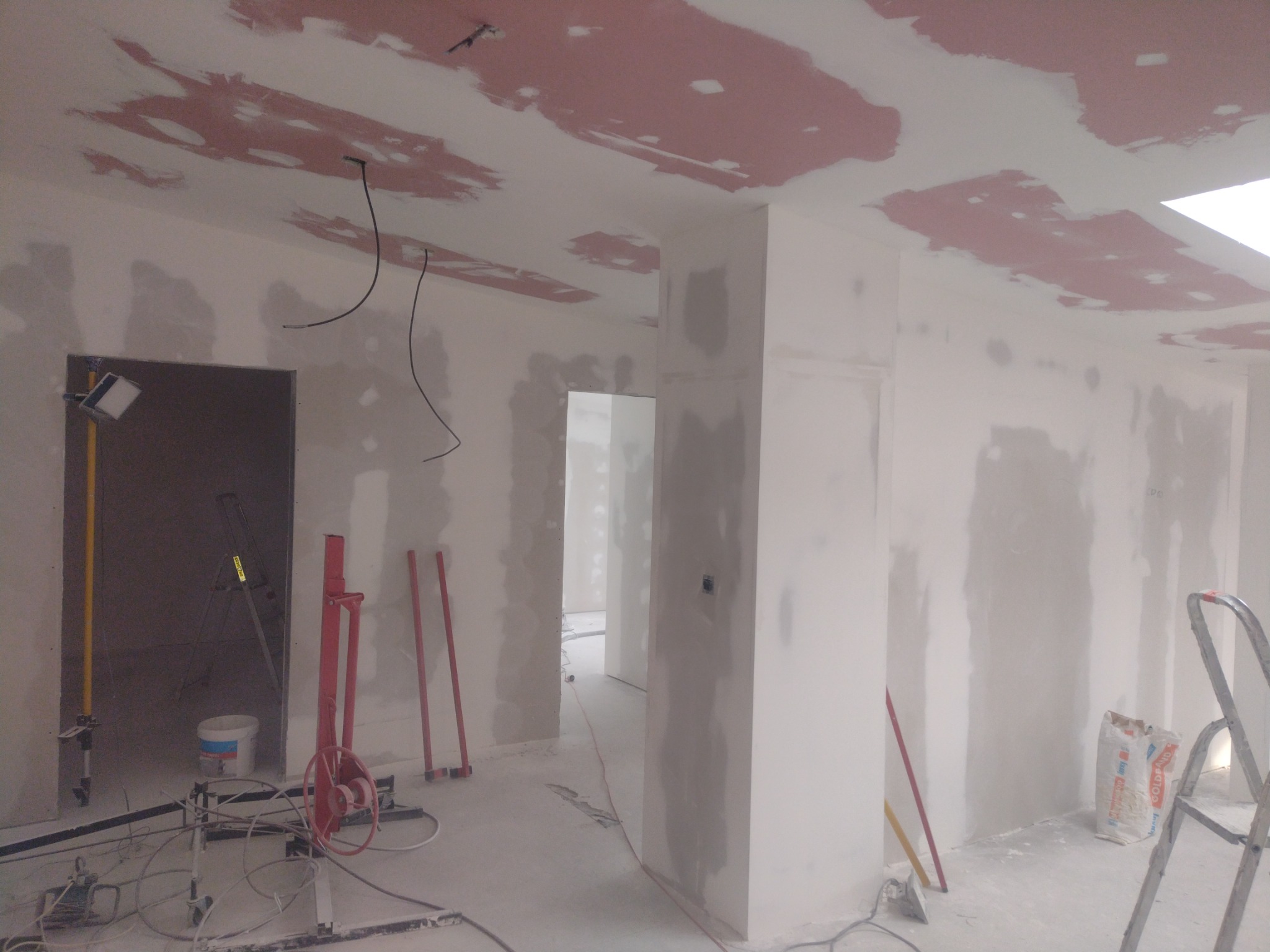 Wnętrze w trakcie prac wykończeniowych: ściany i sufit pokryte warstwą szpachli, widoczne narzędzia, drabina, lampa robocza i worki z zaprawą.