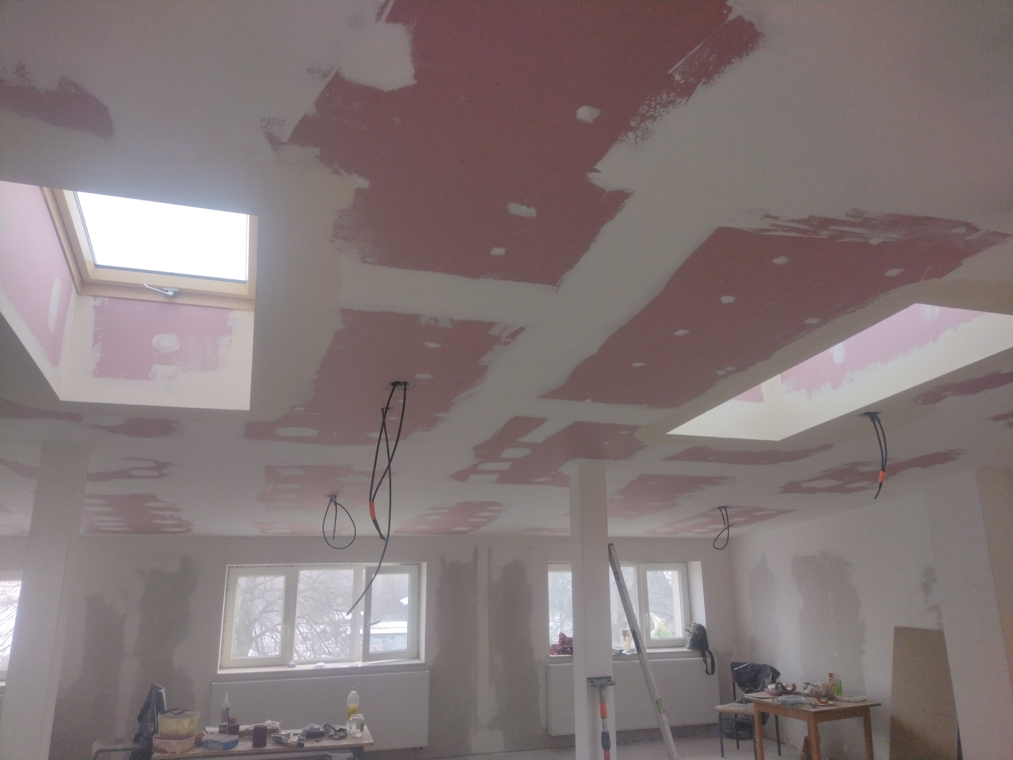 Pokój w trakcie remontu, widoczne ślady po gładzi na suficie i ścianach, dwa okna dachowe, kable zwisające z sufitu, narzędzia i materiały budowlane na stole.