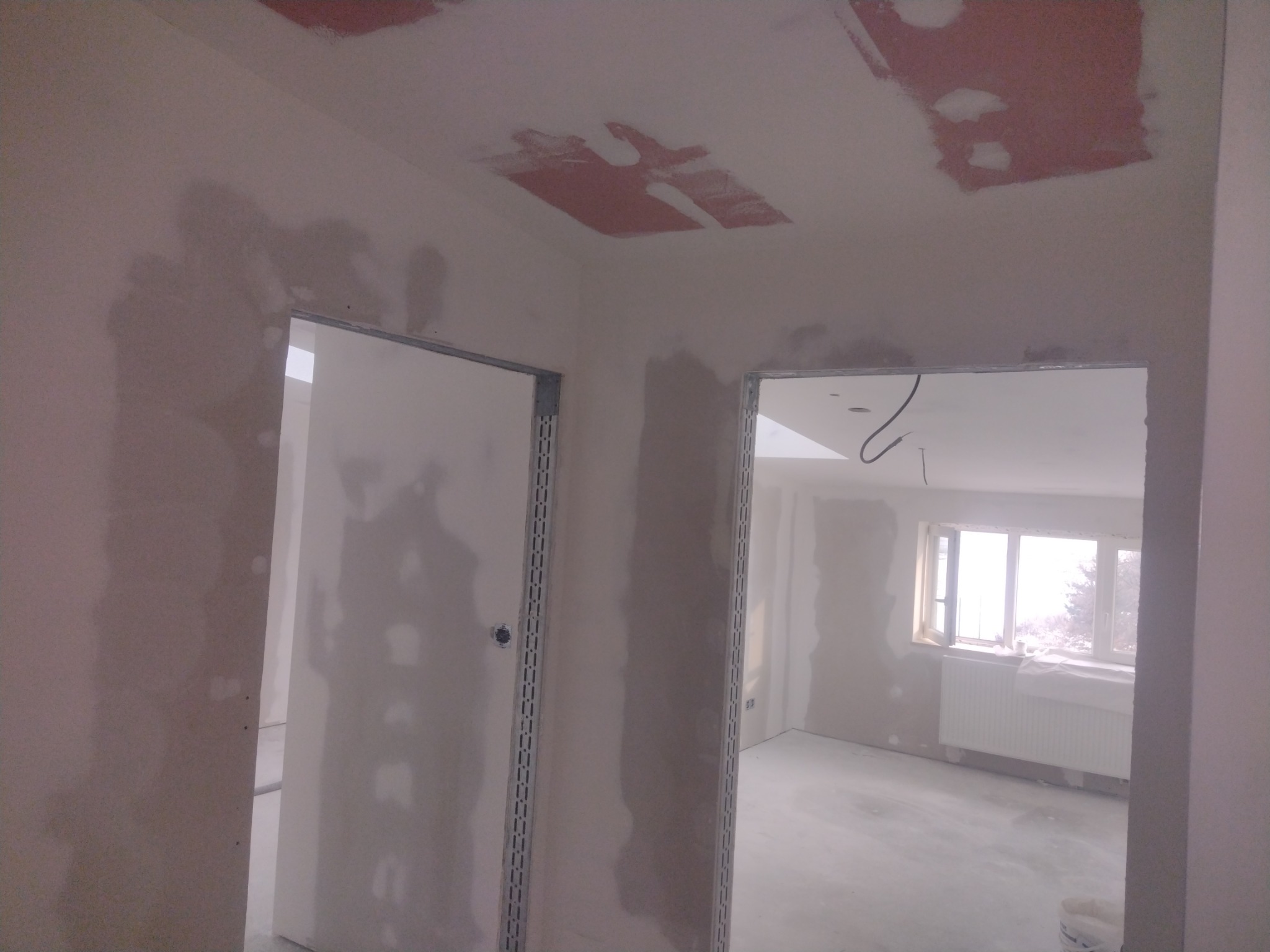 Wnętrze w trakcie remontu: ściany i sufit po nałożeniu gładzi, widoczne metalowe ościeżnice drzwiowe, fragment grzejnika pod oknem.