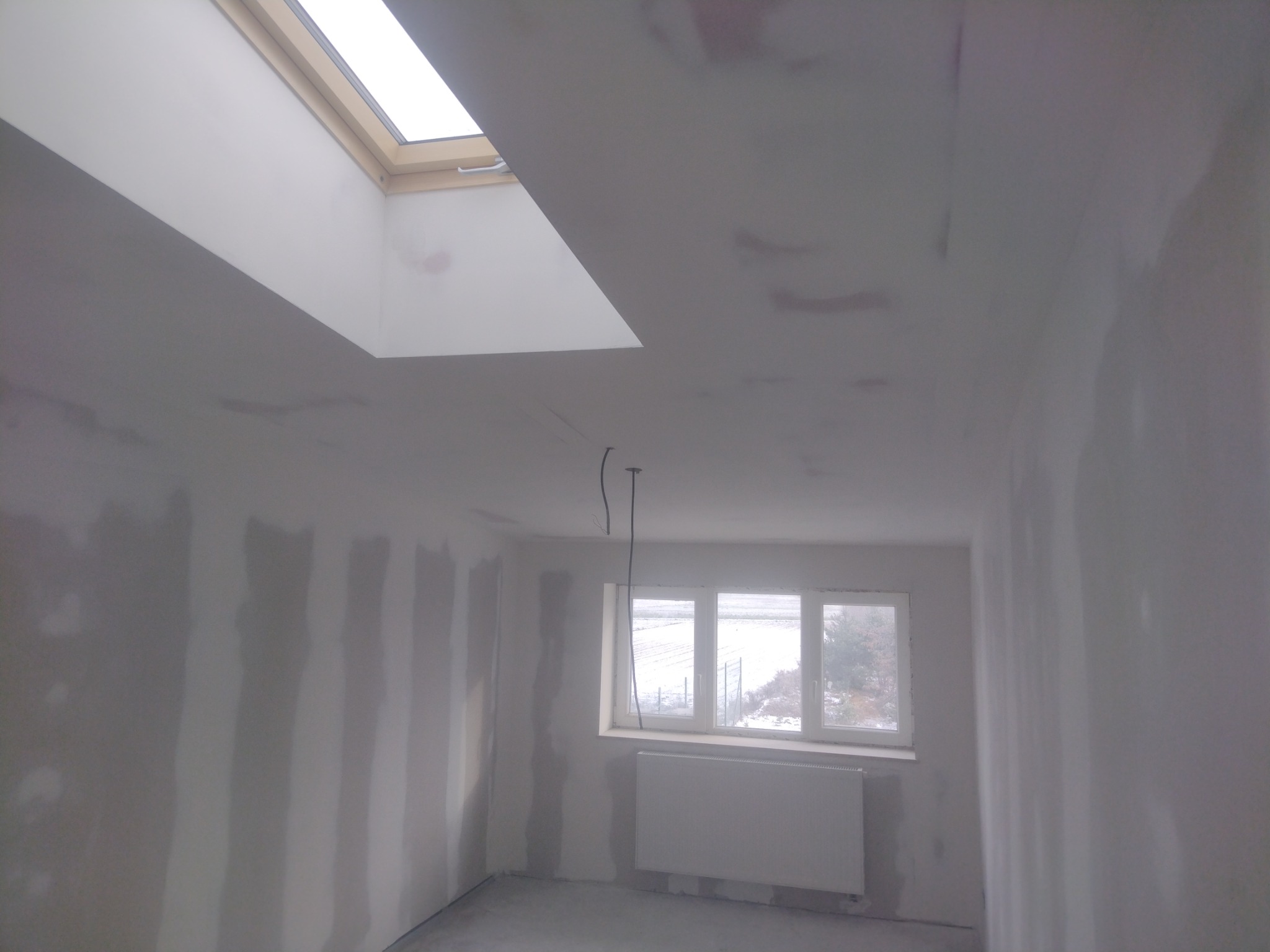 Pokój w trakcie remontu: ściany po nałożeniu gładzi, widoczne ślady szpachlowania, okno z widokiem na zimowy krajobraz, sufit z oknem dachowym.