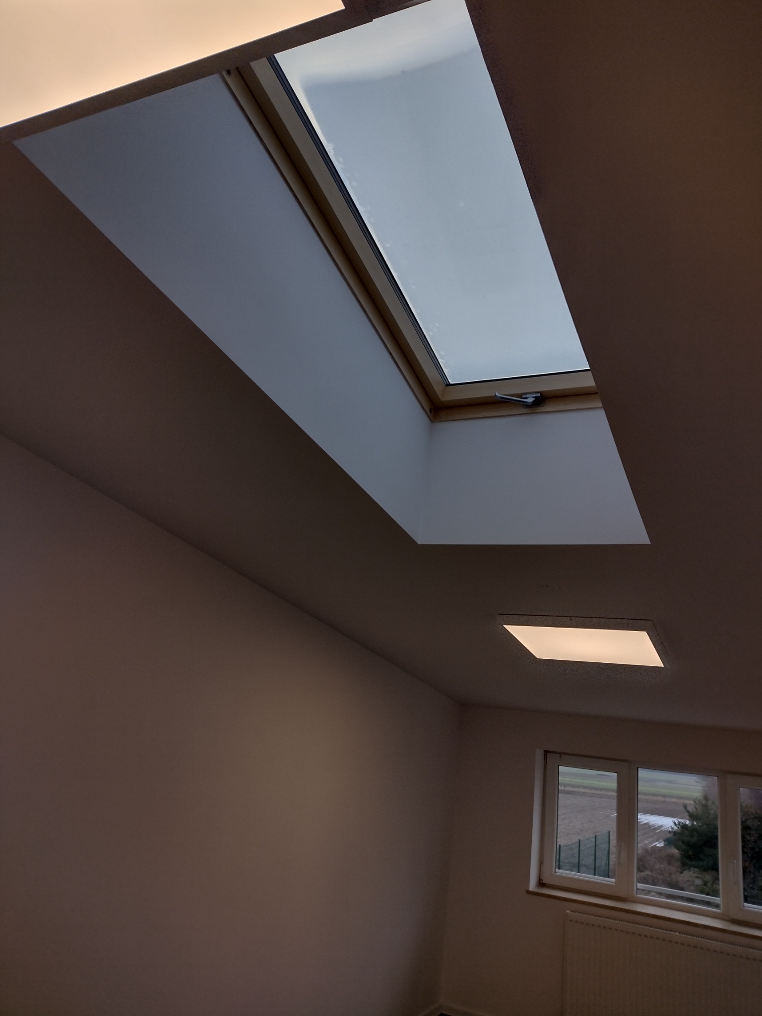 Wnętrze pokoju z oknem dachowym z drewnianą ramą i widocznym mechanizmem otwierania, dodatkowo widoczne okno na ścianie i grzejnik.