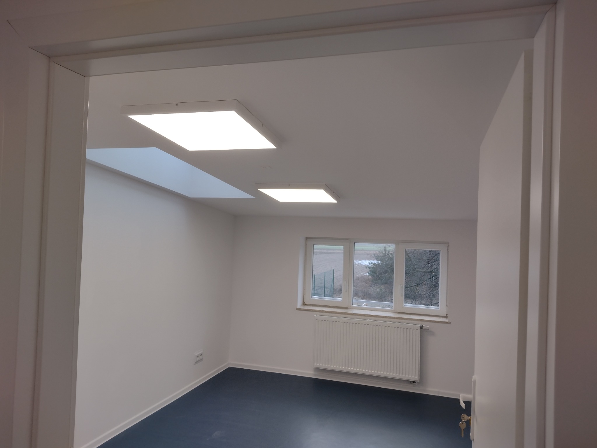 Pomalowane na biało wnętrze z niebieską podłogą, widok zza futryny, okno z grzejnikiem, dwa kwadratowe panele LED na suficie i świetlik.