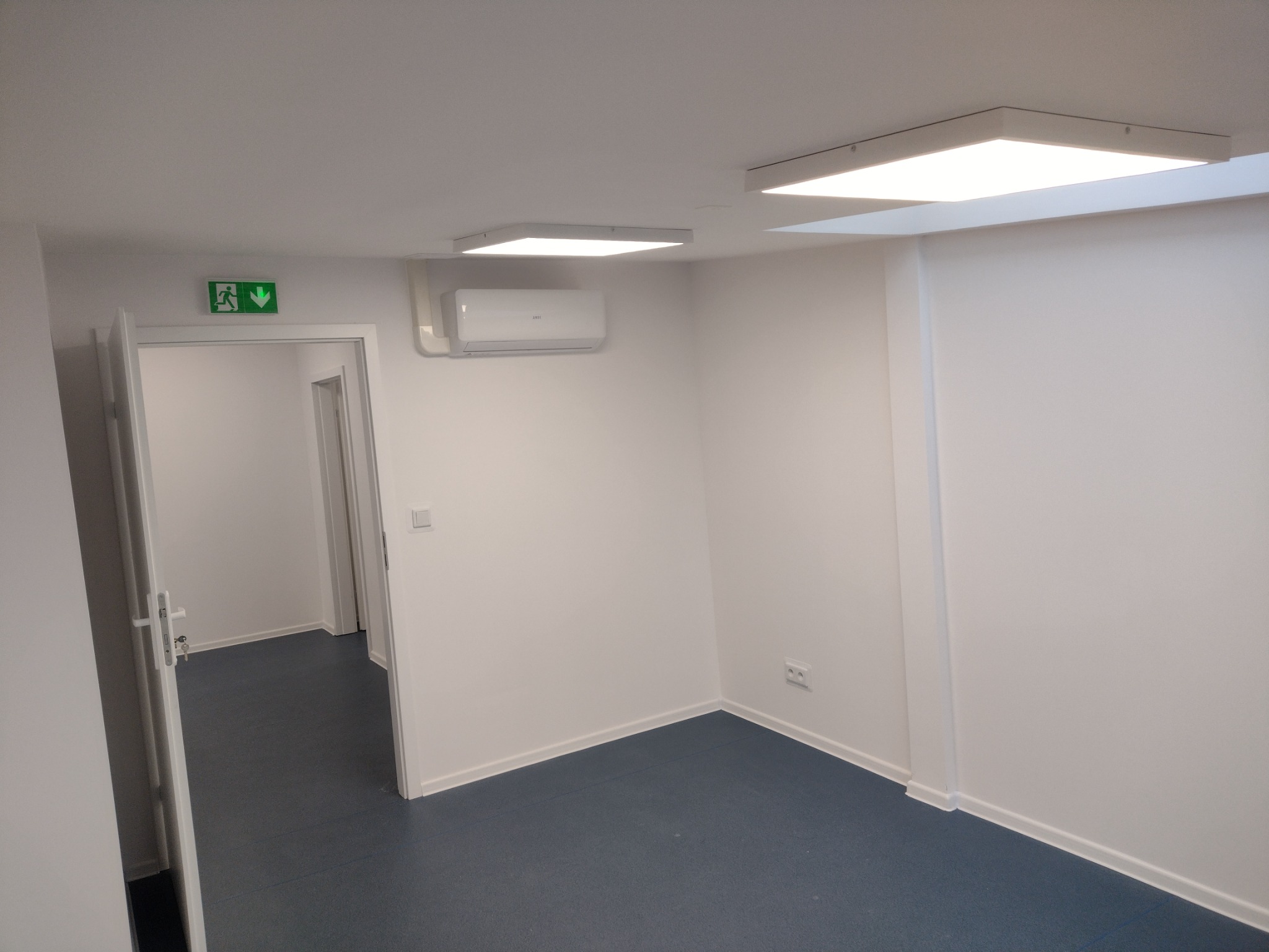 Wnętrze biura w Warszawie po remoncie: białe ściany, sufit z wbudowanym oświetleniem LED, klimatyzacja, szara podłoga i otwarte drzwi z zielonym znakiem ewakuacyjnym.