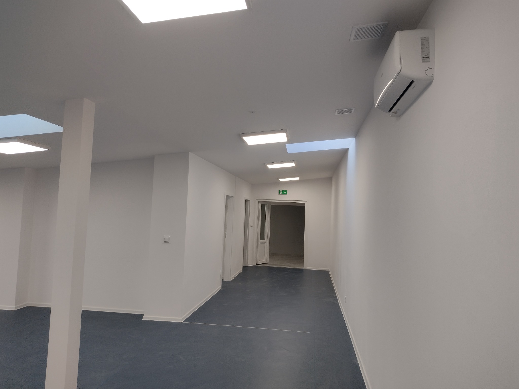 Wnętrze biura po remoncie: białe ściany, szara podłoga, klimatyzacja na ścianie, nowoczesne oświetlenie sufitowe i widoczne drzwi do kolejnego pomieszczenia.