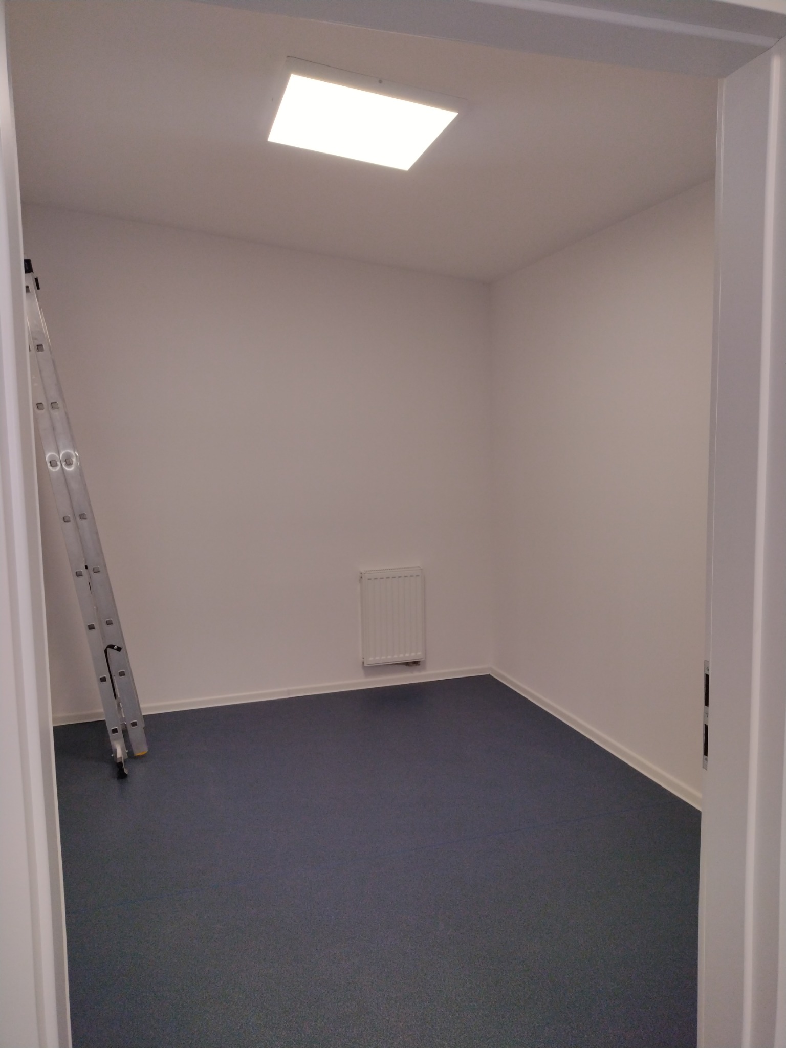 Pomalowane na biało puste pomieszczenie z szarą podłogą, drabina oparta o ścianę, widoczny grzejnik i sufitowe oświetlenie LED.