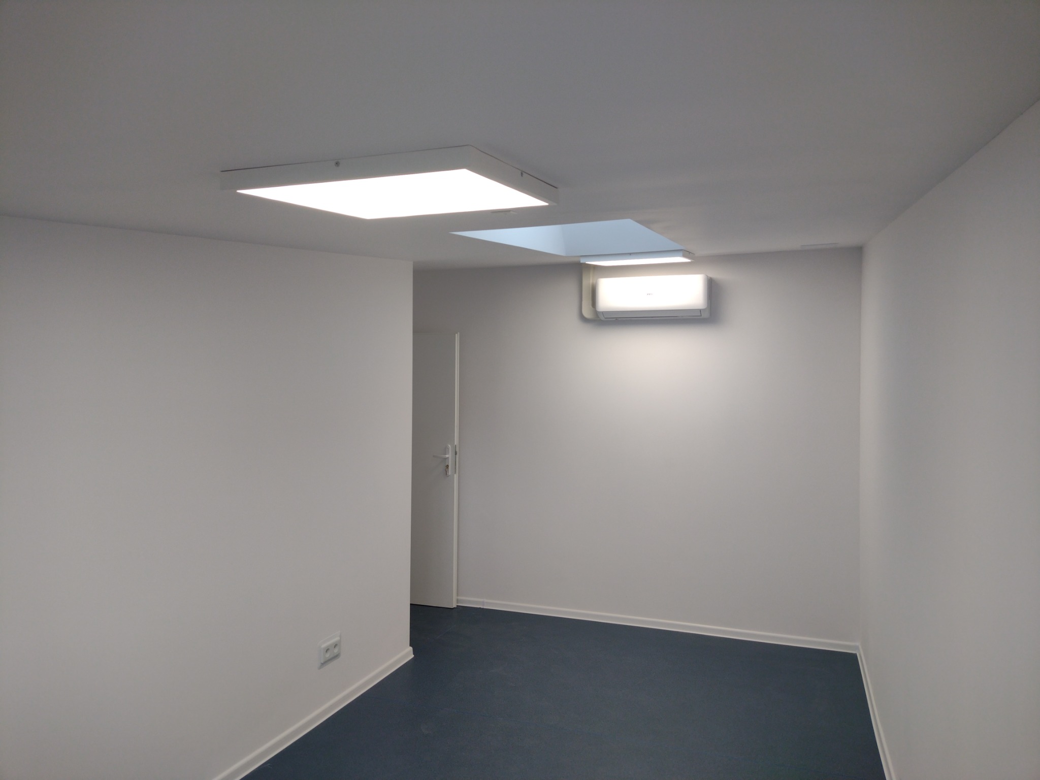 Puste, odnowione pomieszczenie biurowe z białymi ścianami, niebieską podłogą, dwoma panelami LED w suficie i klimatyzatorem na ścianie. Widoczne gniazdko elektryczne przy podłodze.